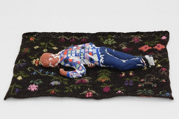 Close up of a work by Ellen Lesperance. Shows a sculpture of a person in a multicolored outfit laying face down on a textile rug with a flower pattern on a dark background. 