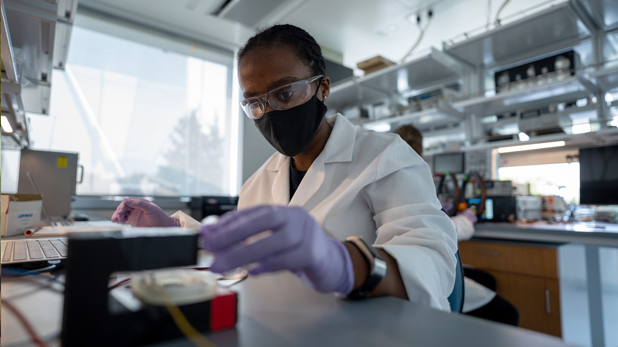 A researcher in a lab coat and gloves works at a lab bench.