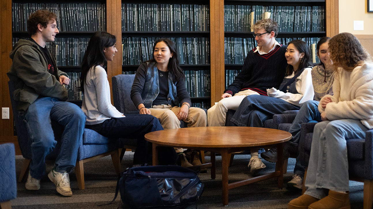 group of students seated in lounge chairs around low table in the Chapman Library in front of shelved theses, talking to one another