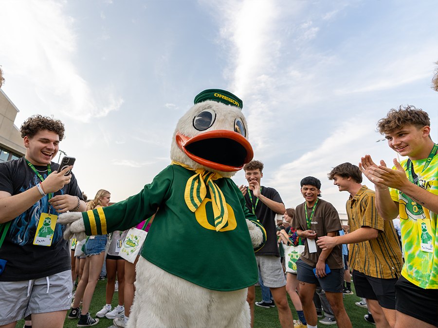 A duck mascot greets a crowd as people clap and use their phones.