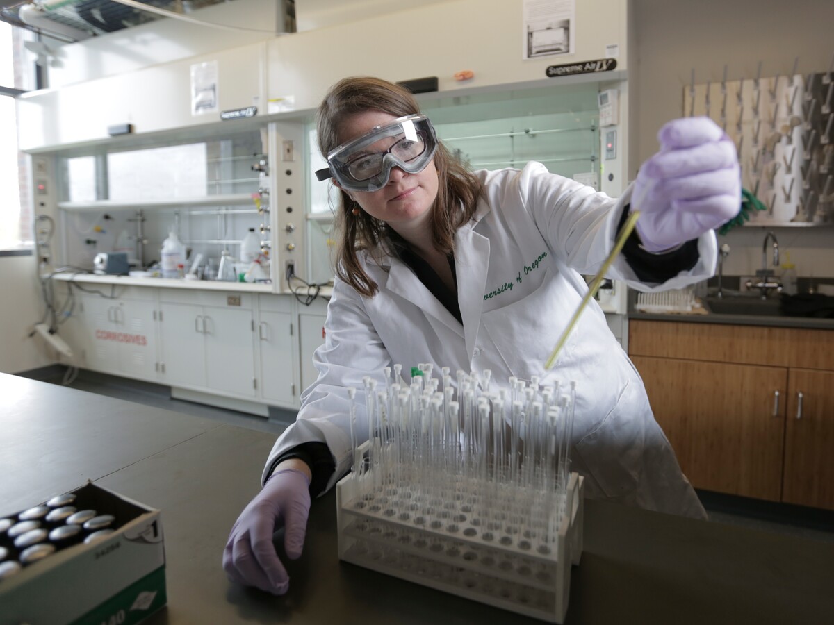 A chemist in a University of Oregon lab coat measures a sample at a fume hood.