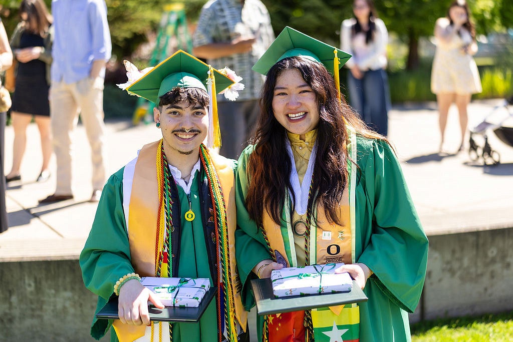 Two smiling graduates in green University of Oregon caps and gowns pose outdoors, holding ribbon-wrapped diploma cases and wearing multiple honor stoles.