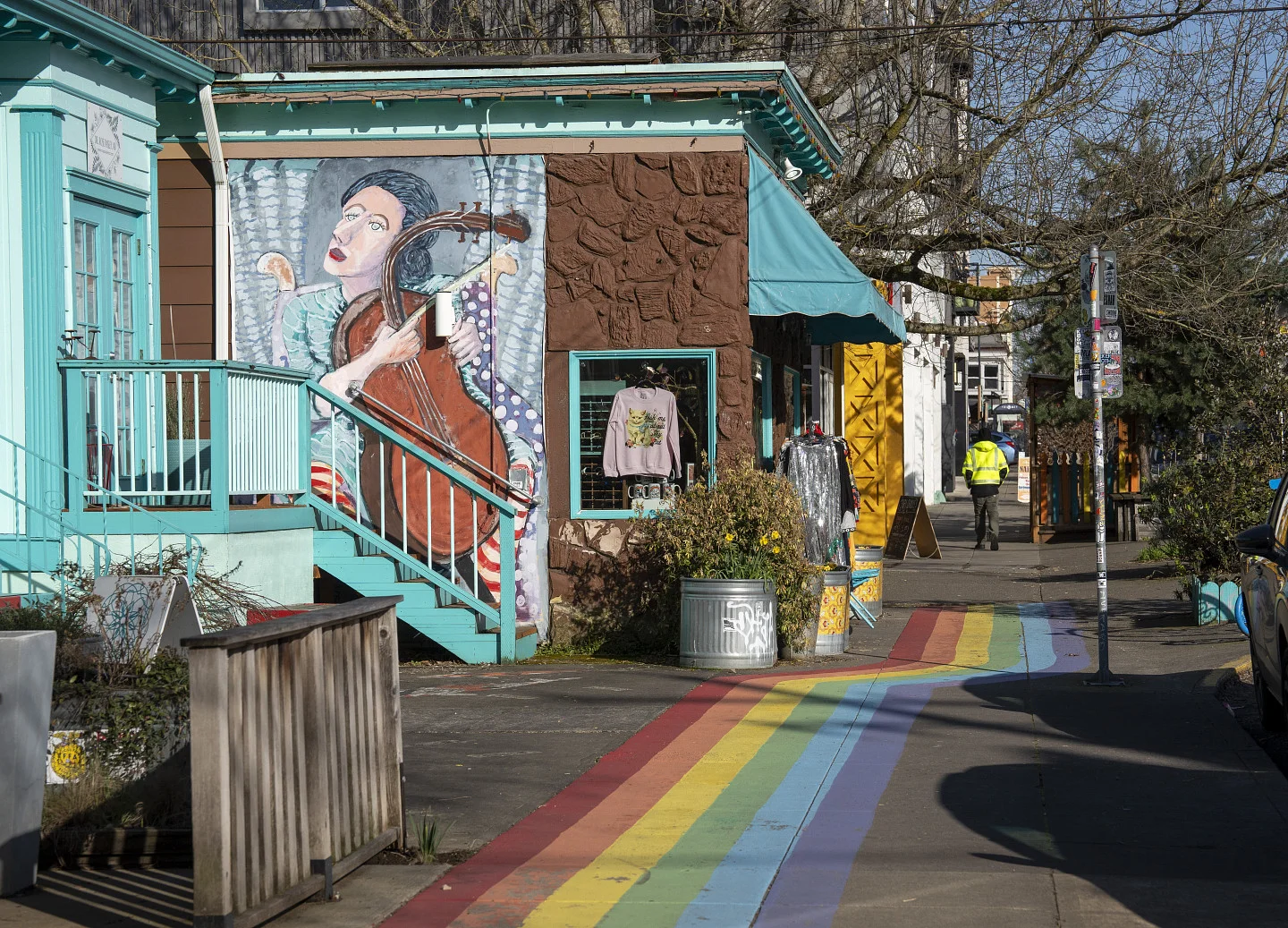A colorful sidewalk painted like a rainbow runs past a building with a mural of a musician playing cello; a person in a yellow jacket walks in the distance.