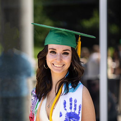 Smiling woman with shoulder length, dark brown hair wearing a green graduate cap and gold tassel, with a yellow-green gradient background.