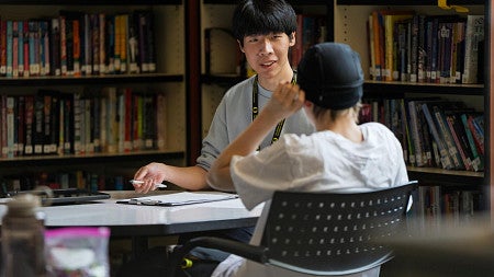 A Ballmer Institute student wearing a University of Oregon lanyard meets with a young person at a table in a school library.
