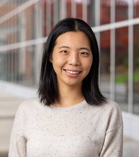 Headshot of a woman with straight black hair smiling outdoors in front of a modern campus building