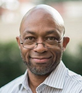 A man with glasses and a light, striped shirt poses in front of a green background