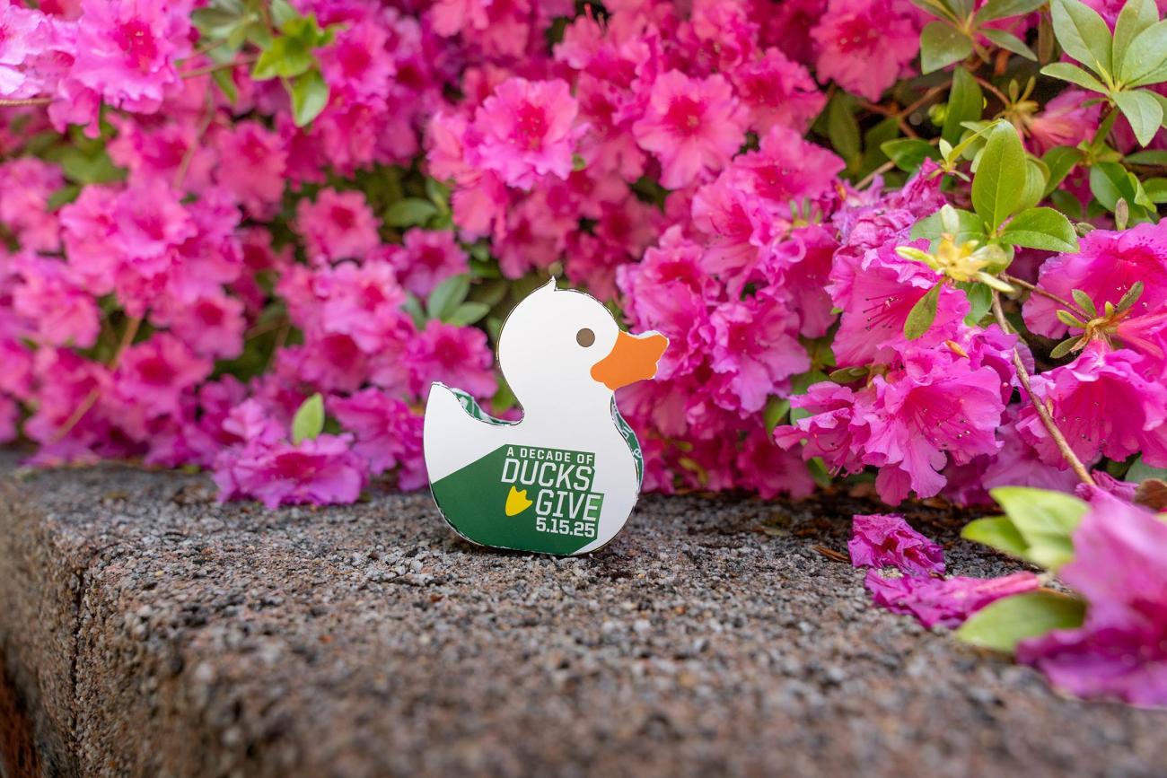 A paper DuckFunder Duck sitting on ledge in front of some flowers.