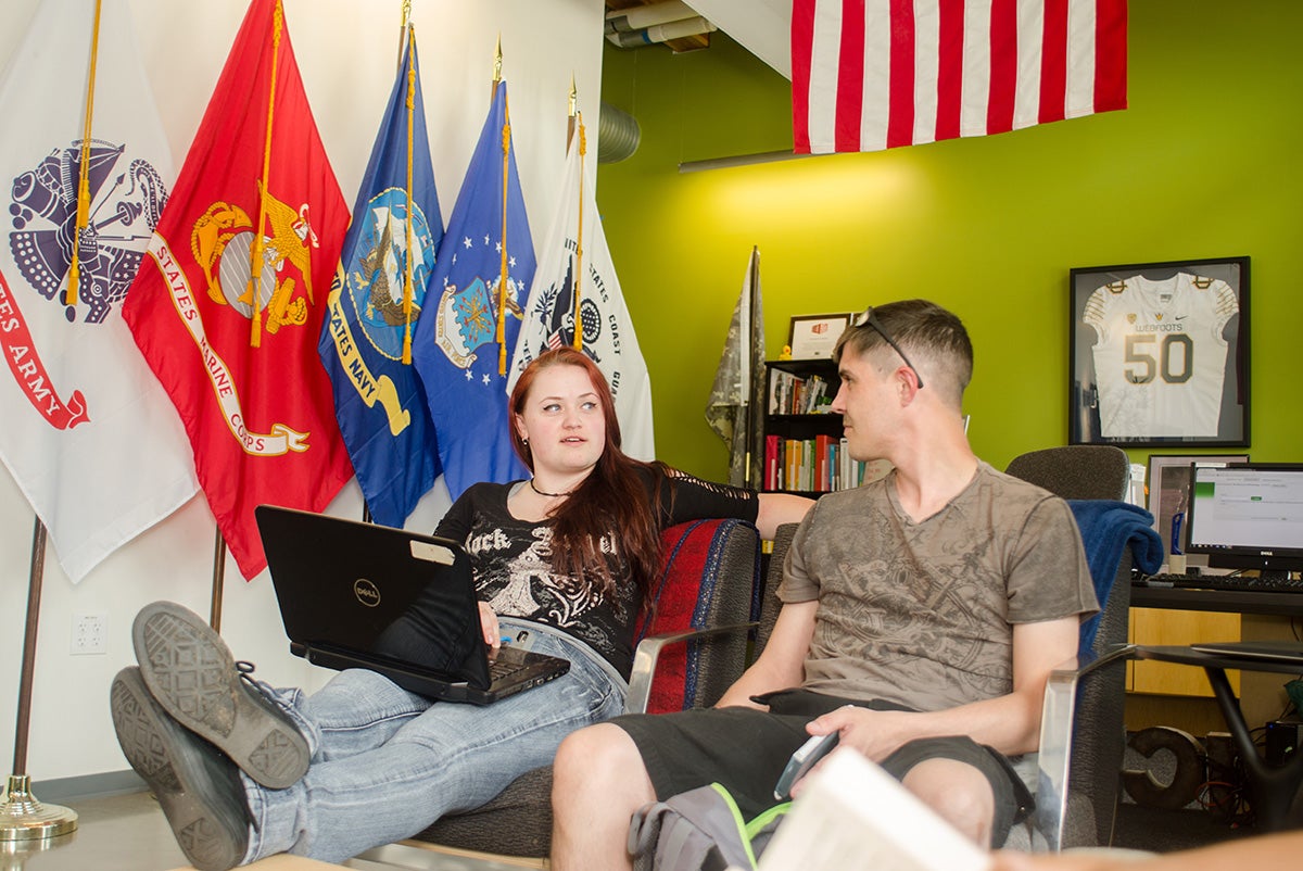 A woman and man sit in lounge chairs with a wall of military flags in the background