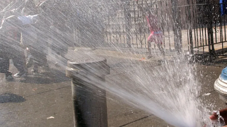 A person uses their hand to direct water from a fire hydrant toward a group of people standing on a sidewalk trying to cool down in the harsh heat