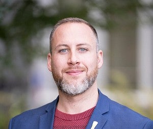 Man with grey and brown beard smiling. Wearing blue collared shirt over a red shirt.