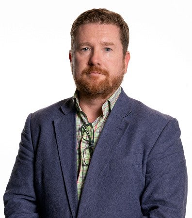 Man with a beard and mustache wearing a plaid green shirt and blue blazer.