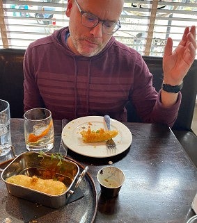 Dean Mundy photo wearing a hooded sweatshirt and eating fried pickle at a restaurant table with hand in air
