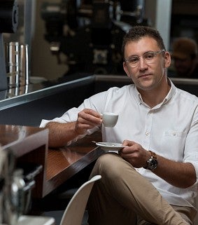 Man with glasses in white shirt sitting in a coffee shop drinking a cup of coffee