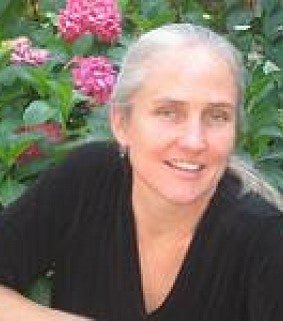 Smiling woman with grey hair in black blouse. Background is a bush with pink flowers.