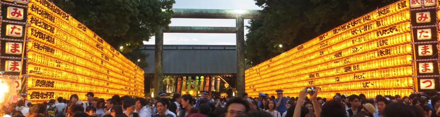 A dense crowd gathers at a festival entrance lined with glowing yellow lanterns covered in writing, leading toward a large gate framed by trees at dusk. 