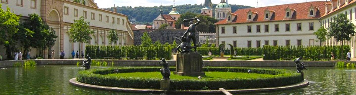  A green garden, with a fountain in the middle surrounded by European style buildings