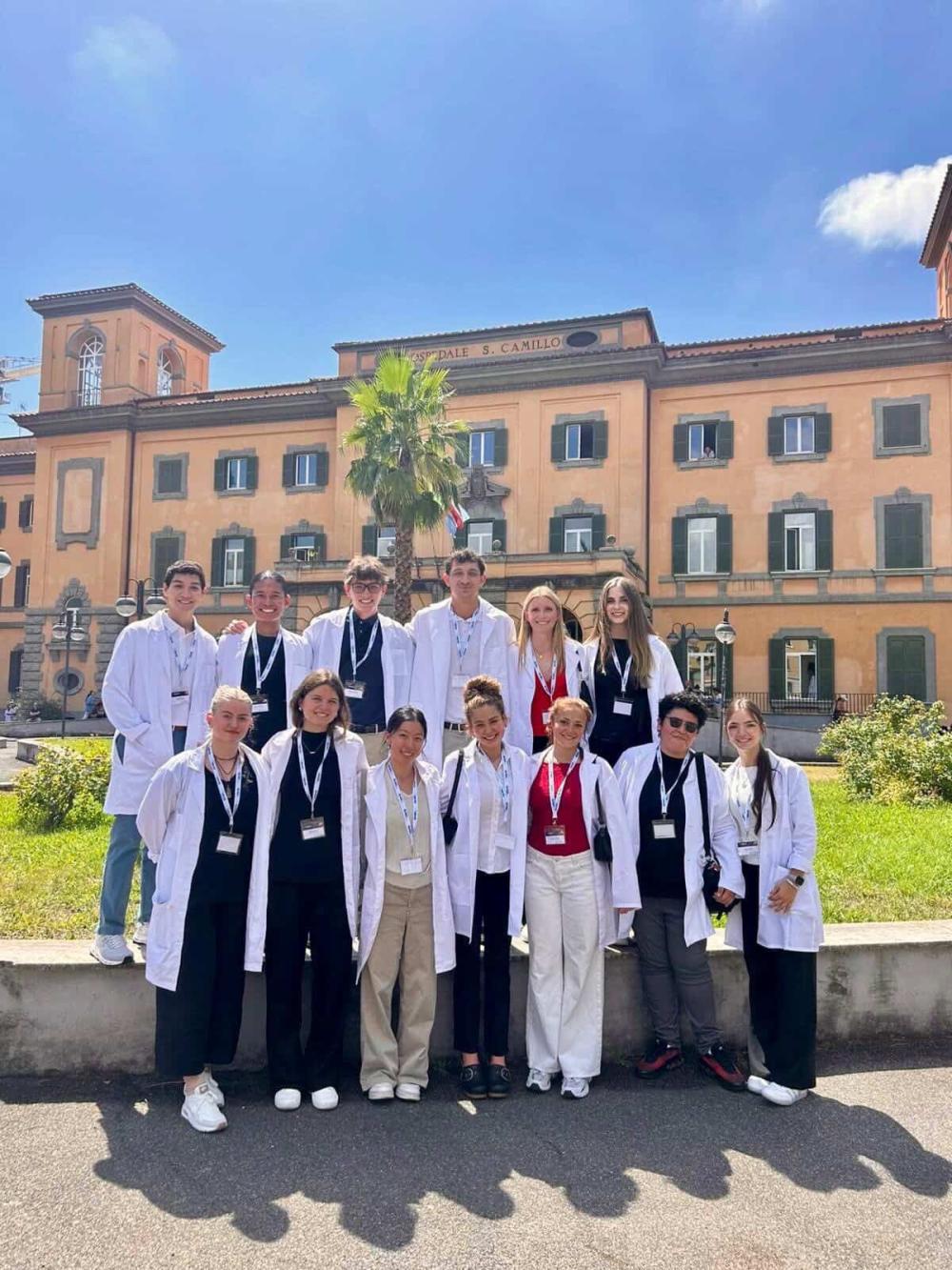 Cohort of premed students smiling outside an Italian hospital wearing lab coats and name tags