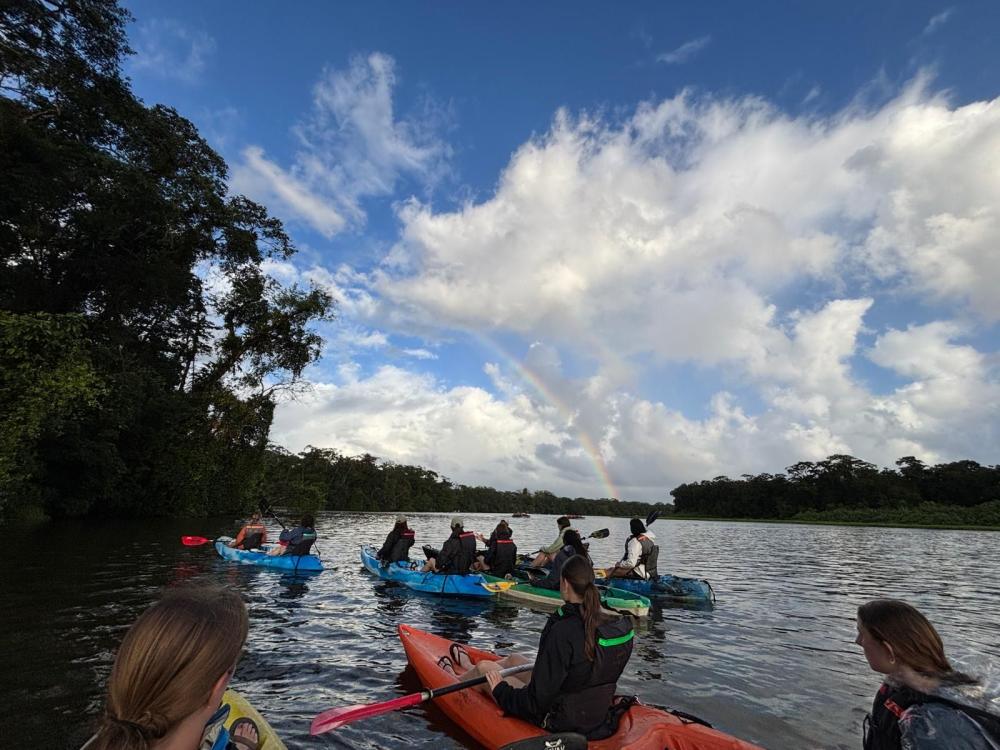About a dozen students kayaking in Costa Rica on a partly cloudy day with a rainbow in the skyline.