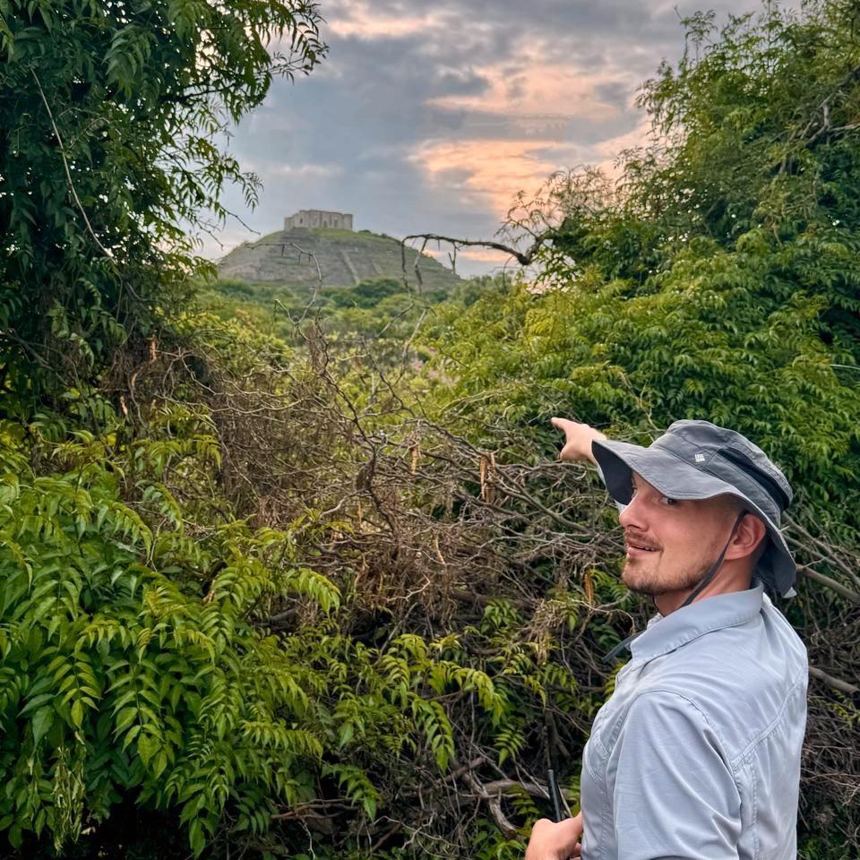 A person in a sun hat points at a mesoamerican pyramid
