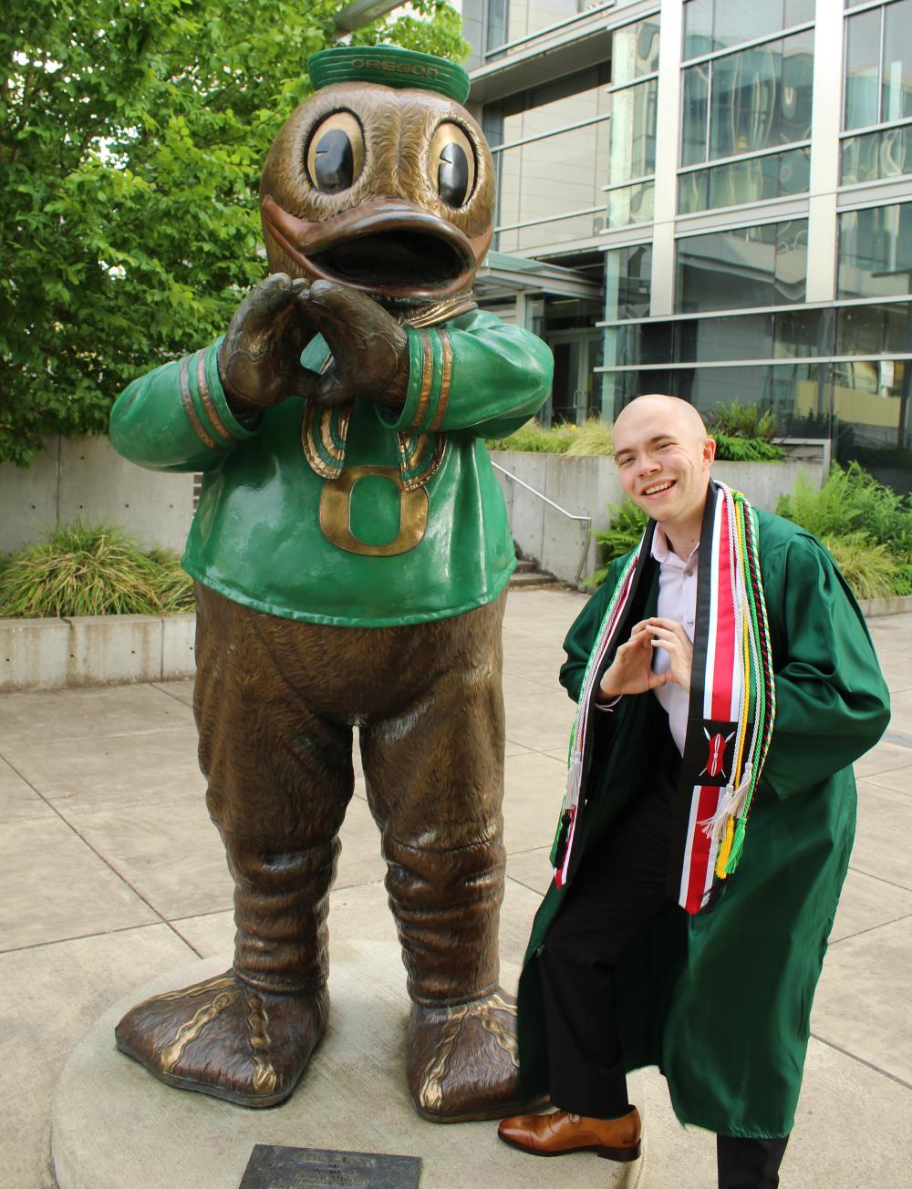 College student wearing green graduation robe posing with UO mascot statue