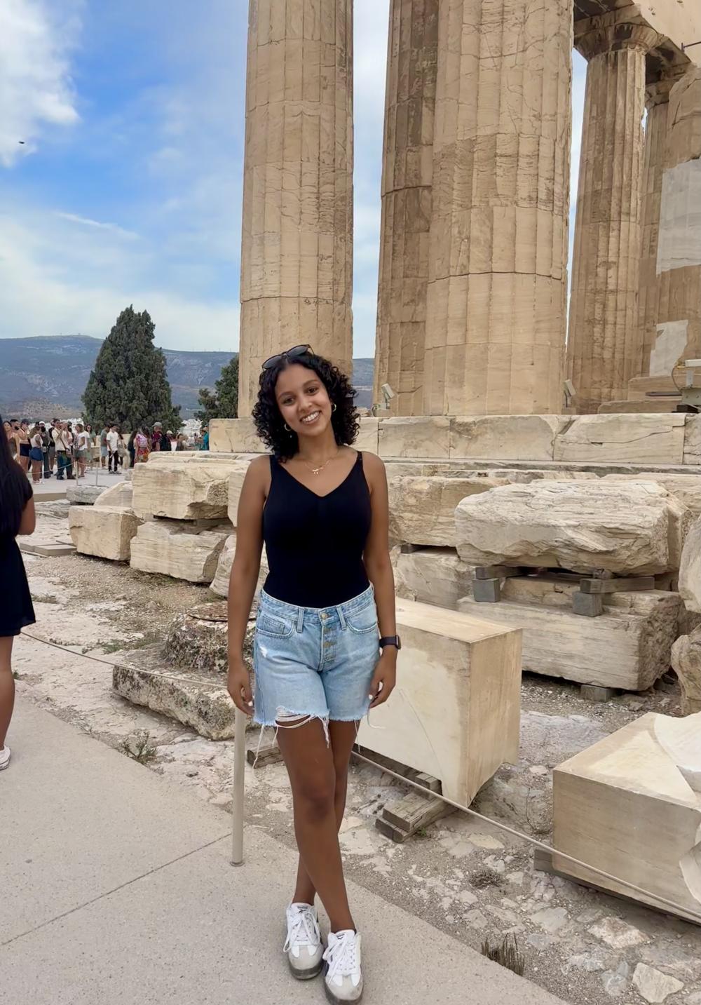 College-aged woman standing by the columns of the Parthenon in Athens, smiling at the camera.