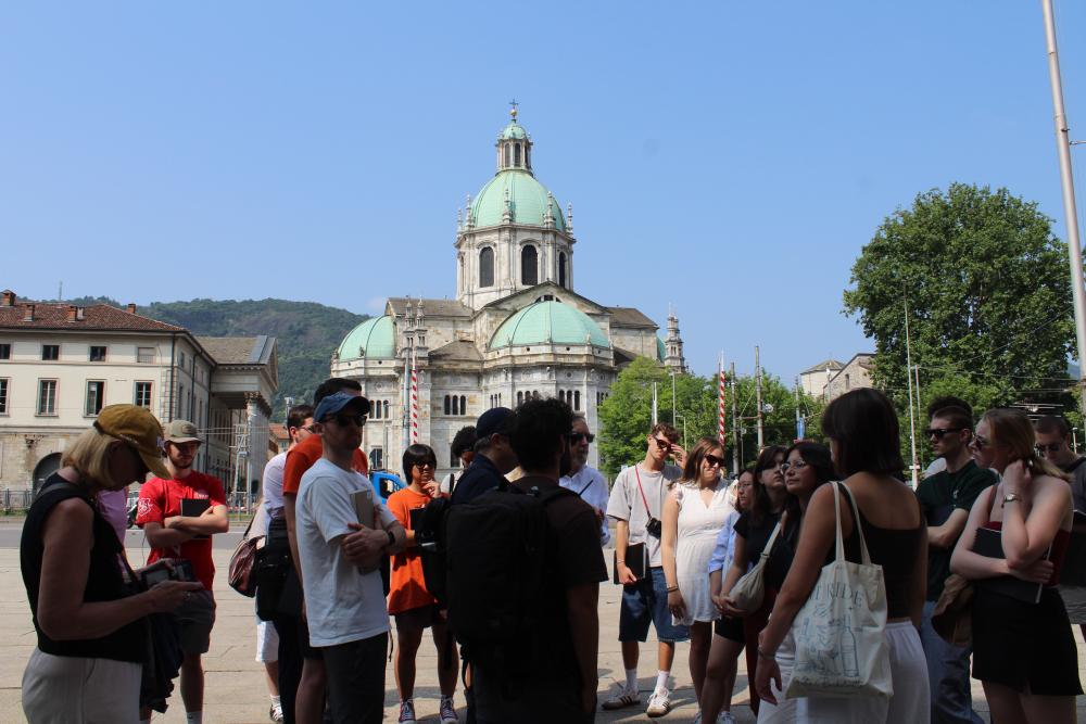 Students stand outside near a promenade of buildings, focused on one speaker