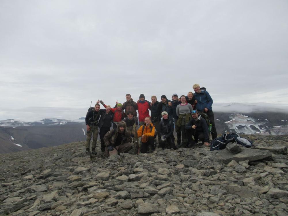 a group of students stand on a hilltop of rocks; snow and clouds are in the background