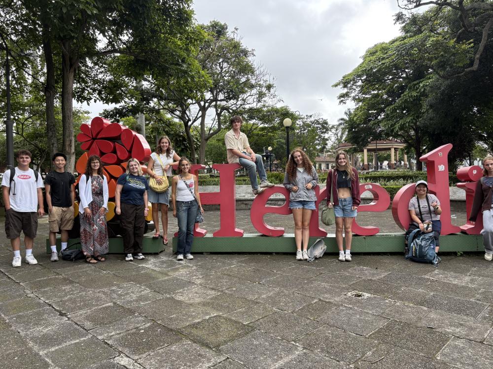 Ten people stand around a large red sign that says "Heredia"