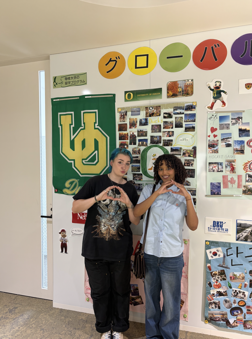 Sara and a friend standing in front of a wall with UO posters and memorabilia, throwing their "O"