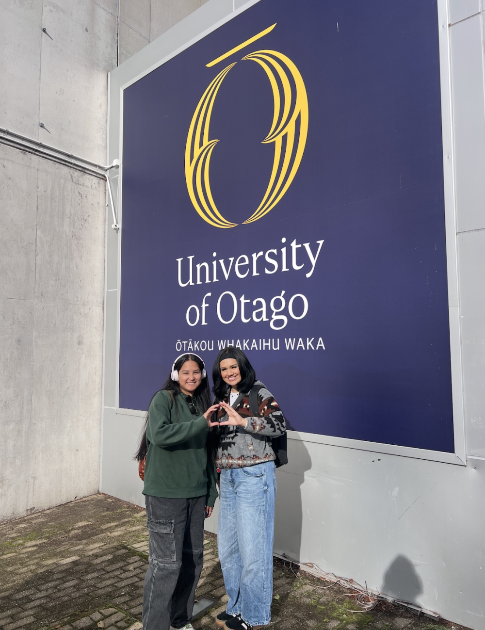 Two people pose, throwing their "O" in front of a University of Otago sign in the background