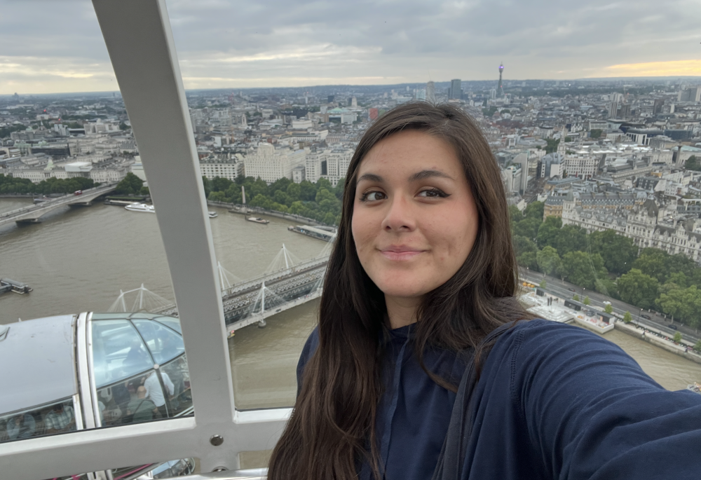 Nathalie poses for a selfie in a clear ferris wheel, with the city skyline of London below