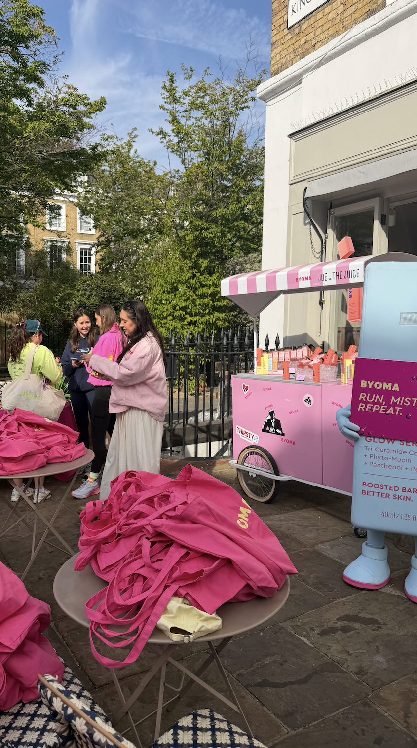 a group of people is in the background, a "Joe and the Juice" pink brand activation cart is beside them; there are pink tote bags in stacks throughout the photo