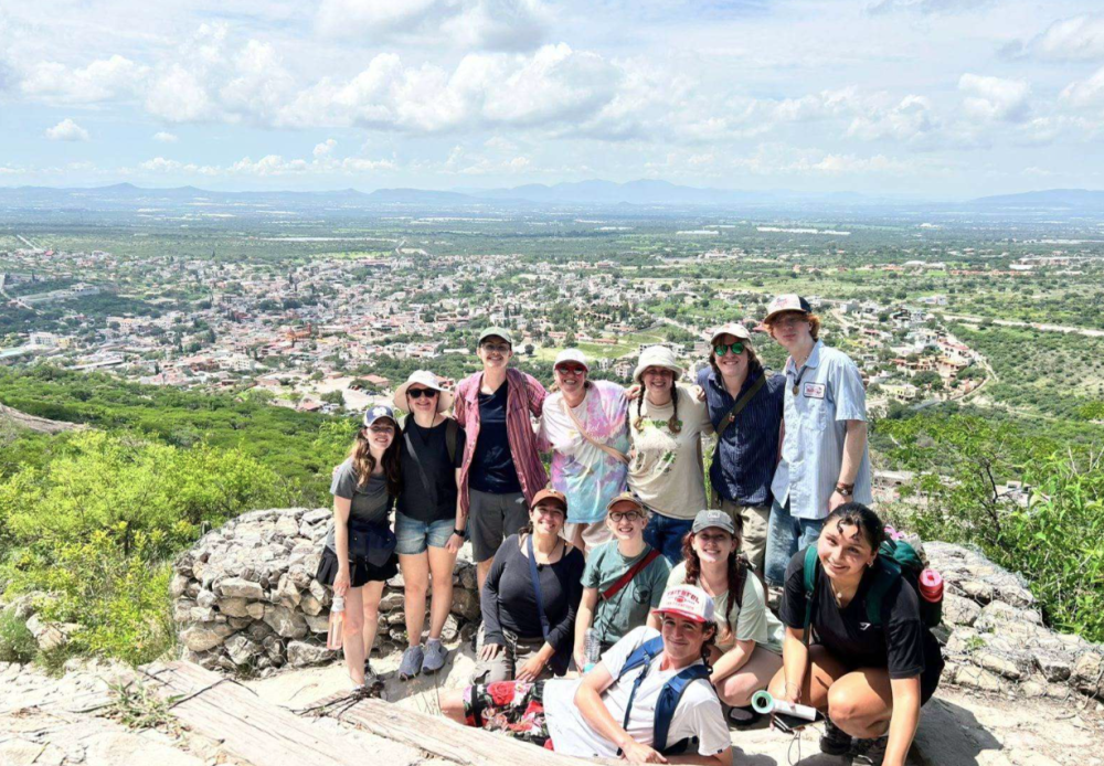 a group of students in hiking gear pose at the top of a rock with the city in the background