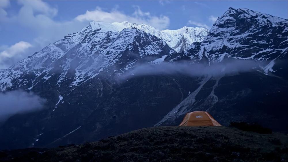 Landscape shot of a tent pitched at dusk with low hanging clouds and snowy mountains in the background