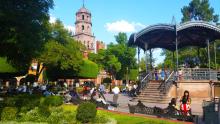 A vibrant public plaza filled with people, featuring a central ornate gazebo and lush green trees.
