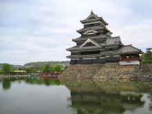A traditional multi-tiered castle with dark roofs rises from a stone base beside a calm moat, reflecting in the water under a pale sky.