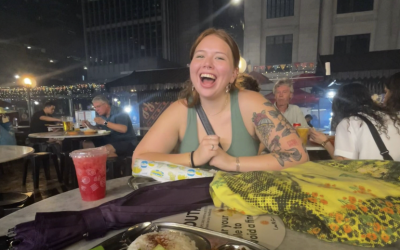 College-aged woman sitting at an outdoor restaurant table at night, smiling with a plate of Singaporean cuisine in front of her.