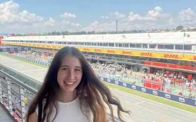 Person smiling at the camera with a Formula 1 race track bustling with activity in the background.