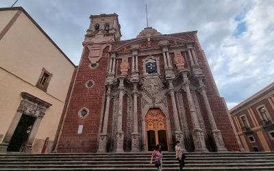 Exterior view of the Templo de San Francisco, an ornate colonial-style church with intricate façade details, under a cloudy sky. People are seen walking on the steps in front.
