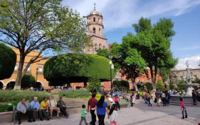 People relaxing and walking in a vibrant public square with lush green trees and a large manicured bush, with a view of a historic church tower under a clear blue sky.