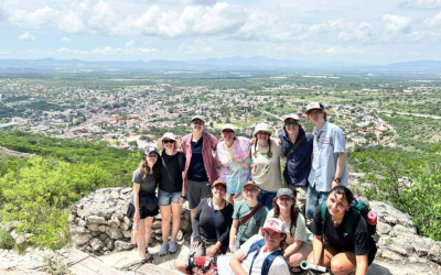 a group of students in hiking gear pose at the top of a rock with the city in the background