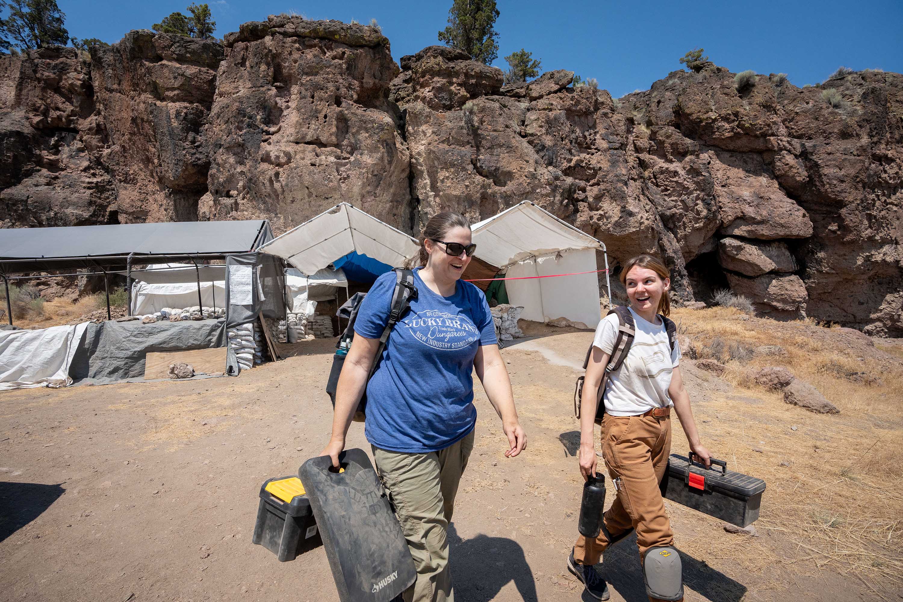Stephanie Alexander and Gabi Gauthier outside walking past tents and carrying gear