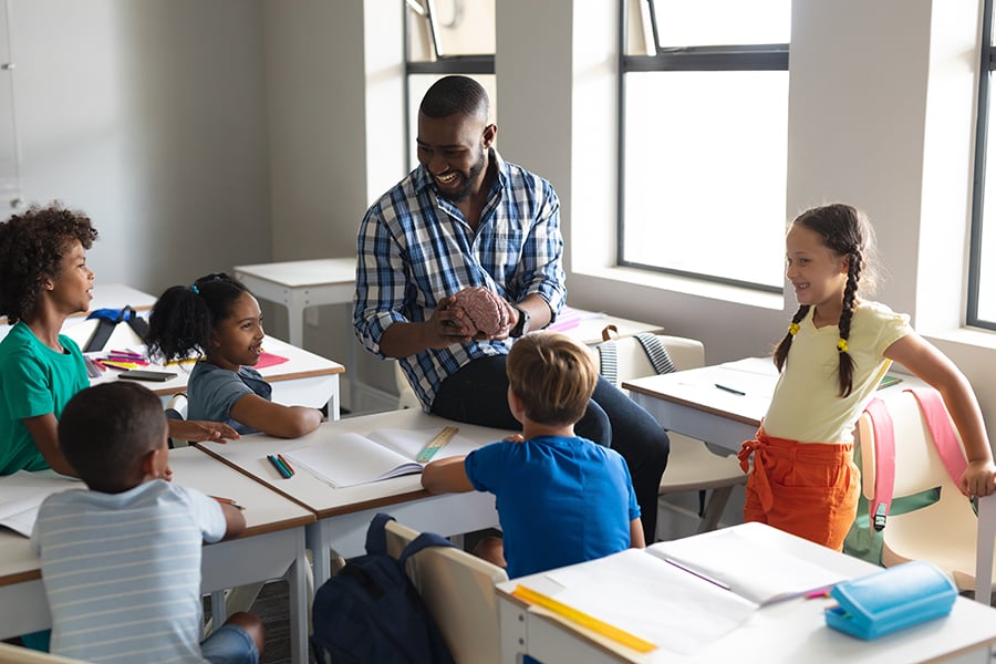 A man seated in a classroom showing a plastic brain to young students