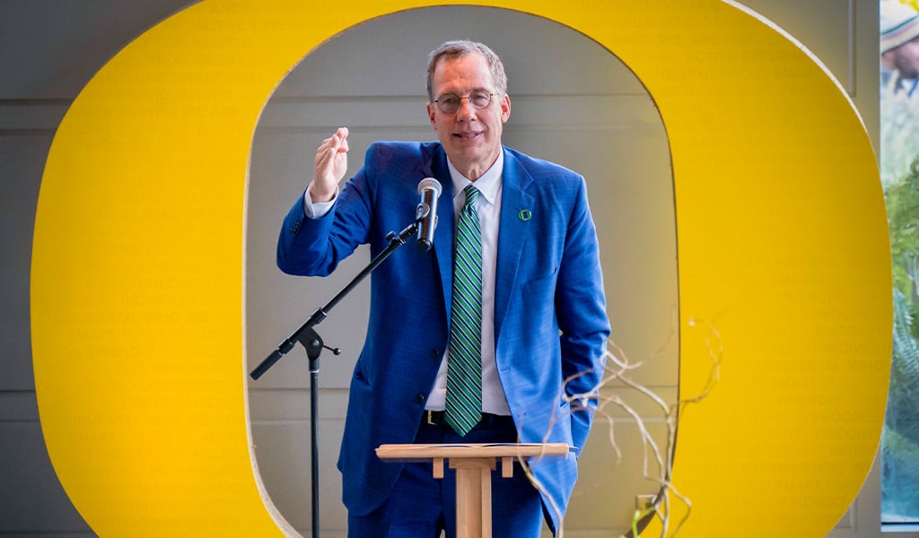 University President speaking to a crowd from a podium in front of a large Oregon O