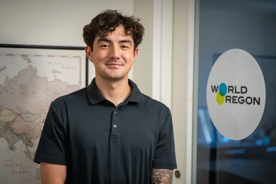 Hunter Eller, intern at World Oregon, stands in front of a world map wearing a black button-up collared short-sleeve shirt. Eller has curly brown hair
