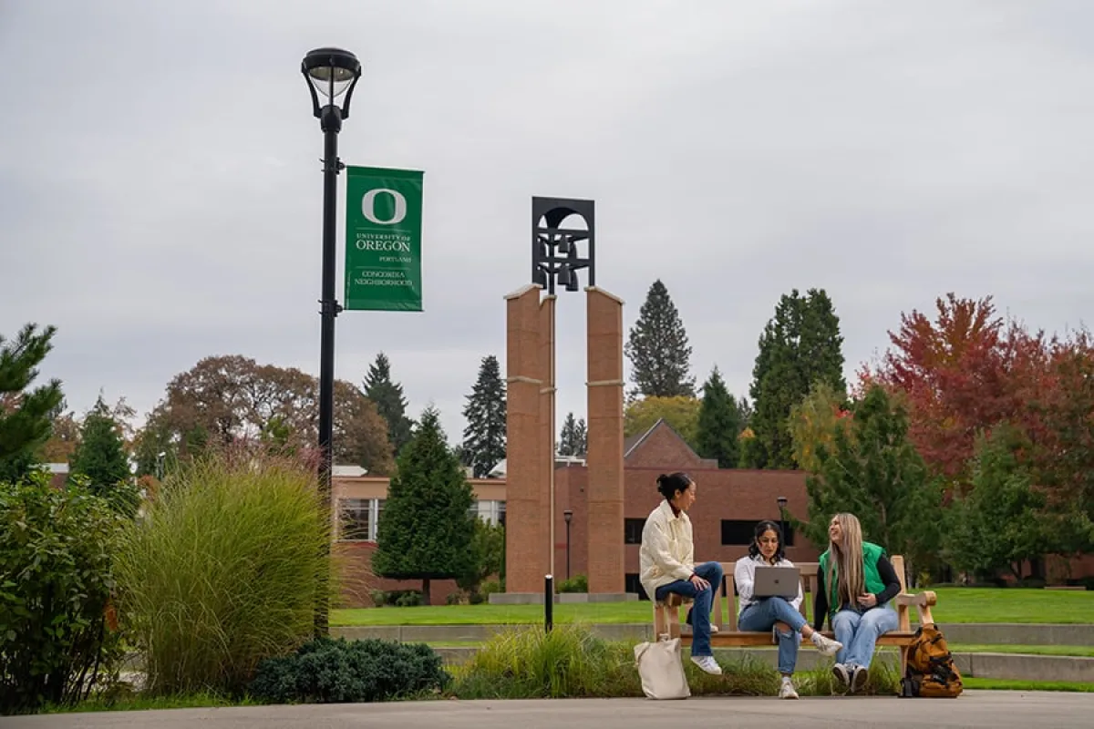Three UO students sit on a bench on campus, chatting and working on a laptop