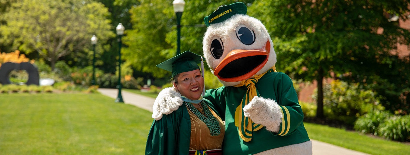 A master's student in graduate regalia poses with the Oregon Duck.