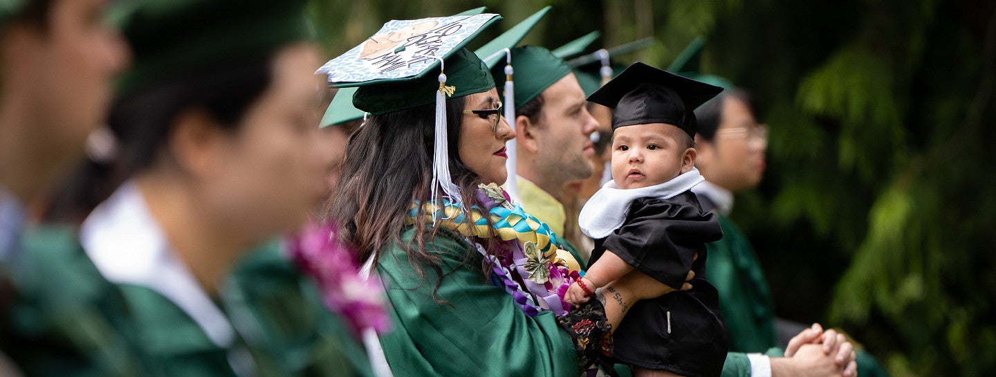 A master's graduate holding their baby, who is dressed in graduation regalia.