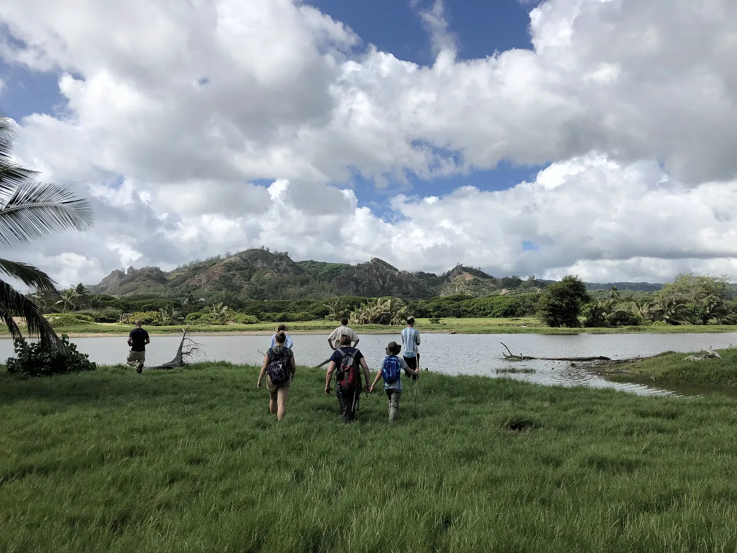 photo of students wearing backpacks hiking in a ocean grass toward a by with mountains in the distance
